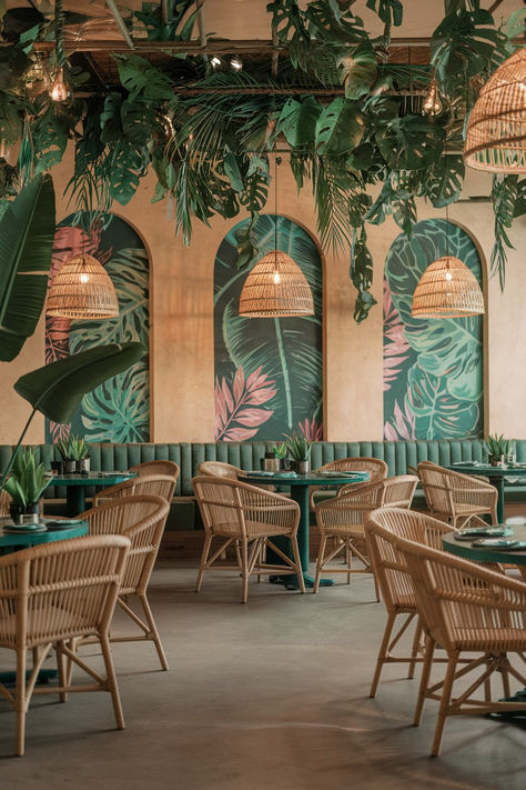 A photo of a tropical bohemian paradise restaurant interior. There are lush green plants hanging from the ceiling and placed in pots around the room. The furniture includes rattan chairs and tables. The walls have woven pendant lights and feature vibrant tropical prints. The room has a warm, earthy tone with a color scheme of emerald green and terra cotta. Emerald Walls, Tropical Restaurant, Restaurant Interior Ideas, Oasis Design, Jungle Vibes, Tropical Boho, Rattan Chairs, Cafe Shop Design, Beach Lounge