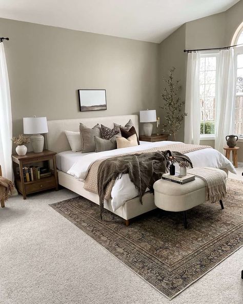 A large modern farmhouse-style bedroom is designed with plush wall-to-wall carpet in a neutral light grey color. There is an antique black and brown carpet centered under the bed for additional interest and comfort. The cream fabric bed is styled with brown and tan blankets and accent pillows, and wooden side tables feature table lamps for a cozy atmosphere...   Image: brittan_allie_home Warm Bedroom, Upholstered Panel Bed, Neutral Bedroom, Grey Carpet, Master Bed, Primary Bedroom, Large Bedroom, Spare Room, Panel Bed