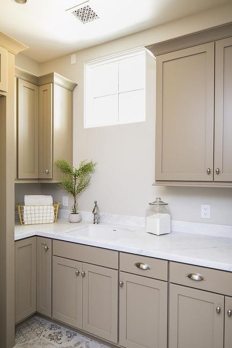 Light brown shaker cabinets accented with satin nickel cup pulls and a white quartz countertop are fitted with a sink matched with a satin nickel faucet located beneath a window flanked by light brown upper cabinets. Brown Painted Cabinets, Light Brown Cabinets, Light Brown Kitchen Cabinets, Light Brown Kitchen, Taupe Kitchen Cabinets, Beige Kitchen Cabinets, White Kitchen Countertops, Taupe Kitchen, Beige Cabinets