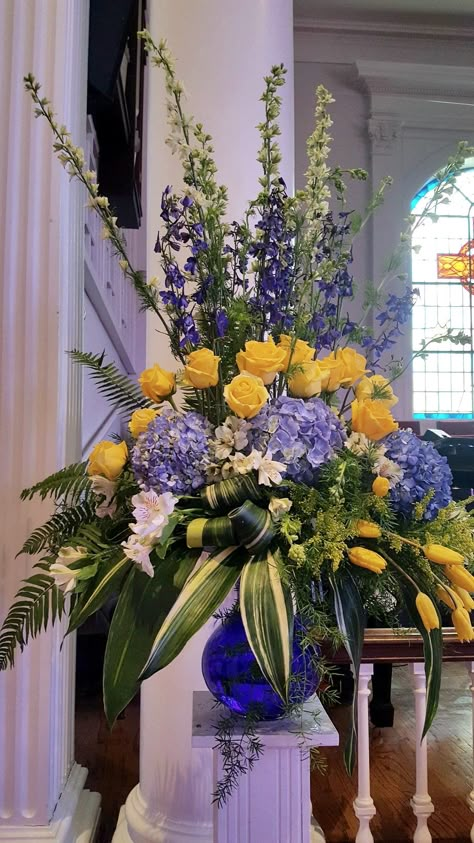 Yellow and blue theme of hydrangeas, yellow roses, yellow,tulips, white larkspur, dark blue delphiniums, lime alstromeria, sword fern, variegated aspedistra, sperengetti fern. https://www.instagram.com/p/CDNSlfQpmNW/?igshid=k5prlwi4adrm Egyptian Floral Arrangement, Blue And White Tall Floral Arrangements, Purple Blue Yellow Flower Arrangement, Easter Florals Diy Church, Blue Yellow Flower Arrangements, Podium Flower Arrangements, Blue And Yellow Flower Arrangements, Easter Flower Arrangements For Church, Cemetery Arrangements