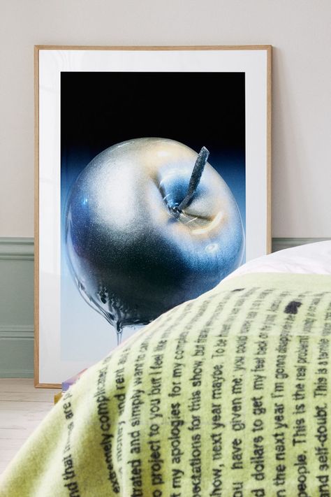 A framed poster of a glittering apple is propped against a wall in a bedroom, adjacent to a bed with white linens. The apple, set against a black background in the poster, has a surreal sparkle that suggests a metallic surface. The duvet is covered with a green throw that has text overlay, which seems like pages of a book projected onto the fabric, creating a cozy yet intellectual atmosphere in the room Apple Poster, Tranquil Bedroom, White Linens, Modern Interior Design, Malaga, A Word, Modern Bedroom, White Linen, Modern Interior