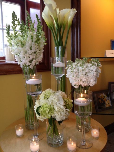 Glass cyldiners designed in white... calla lillies, snap dragons, andstock with a cyldiner of white and mini greenhydrangea surrounded by pedestals with floating candles and votive candles.   Doristhefloristt.com Floating Candles In Cylinder Vases, Cylinder Vase With Floating Candle, Lily Vase Centrepiece, Cylinder Vase Centerpiece Wedding Floating Candles White Flowers, Calla Lillies Wedding, White Lillies In Tall Vase, Hotel Flower Arrangements, 30th Anniversary Parties, Snap Dragons