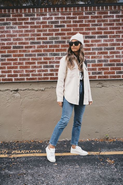 ashley wearing a black graphic tee, off white shacket, mid-wash mom jeans, white sneakers, black square frame sunglasses, and a white beanie cap standing in front of a brick wall Womens Casual Outfits With Sneakers, Cream Shacket Outfit Winter, Charcoal Shacket Outfit, Outfits With Cream Shacket, White Shacket Outfit Fall, Cream Colored Shacket Outfit, Sand Shacket Outfit, Legging And Shacket Outfit, Cream Fleece Shacket Outfit