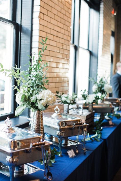 One of the last places couples think to decorate is the buffet table, but sprucing up this space will add some wow factor to your wedding. This couple used a navy linen instead of the typical black, and included some simple vases with a white hydrangea and greenery. Simple, classy, and sure to impress guests. Buffet Dishes Display, Chafing Dish Display Ideas Wedding, Buffet Wedding Decor, Party Buffet Table Set Up, Buffet Centerpiece Ideas, Elegant Buffet Table Ideas Decor, Elegant Wedding Buffet Ideas, Wedding Buffet Ideas Food Stations, Chafing Dish Display Ideas