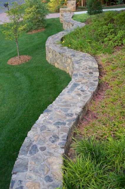 This thick barrier wall is the top of a retainer wall made completely of dark stone. The variation of tone and color make this wall so interesting to look at and it comes completely customizable since you can virtually find all colors of stone out there these days. The way the top of this wall snakes across the garden scene makes the yard seem more put together and clean. Add some flowering plants and trees to the sides of the wall, and you have a custom, beautiful garden. Diy Retaining Wall, Backyard Retaining Walls, Stone Walls Garden, Retaining Wall Ideas, Garden Retaining Wall, Raised Patio, Rock Walls, Landscaping Retaining Walls, Garden Walls