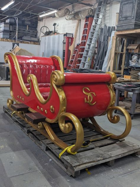 An ornate red sled with gold accents sits on a palette in the shop. Gold letters "SC" are embellished on the front of the sleigh to indicate that it's Santa's. Outdoor Sleigh Decor, Santa’s Sleigh, Santa Sleigh Decoration, Cranberry Farm, Santa Village, Christmas Grotto, Sled Decor, Santa Claus Sleigh, Christmas Story Books