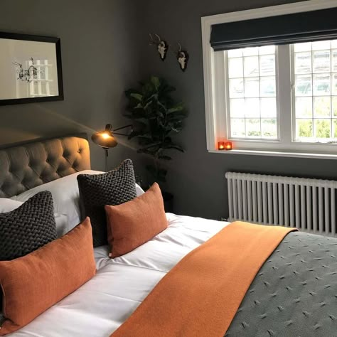 This guest bedroom features a large window that lets natural light into the room, brightening up the dark gray walls. There is a black and white print centered over the tufted grey bedframe. The bed is arranged with crisp white bedding, orange and grey knit accent pillows, and a textured orange and gray blanket. Orange Guest Bedroom, Grey Orange Bedroom, Orange Interior Design, Orange Bedroom Decor, Dark Gray Bedroom, Modern Guest Bedroom, Luxury Home Interior, Home Interior Design Ideas, Orange Rooms