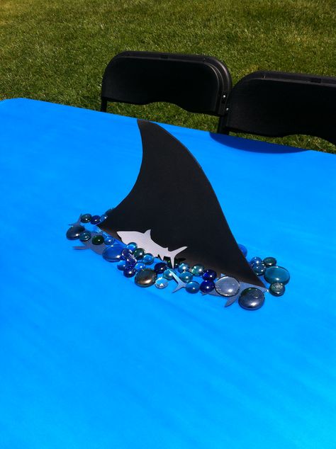 my shark table decor.   foam fin cut out,  cut hole in the table cloth and tape fin to table,  place rocks around the fin to cover edges. Jaws Party, Shark Baby Shower, Shark Week Party, Shark Themed Birthday Party, Shark Cake, Ocean Birthday, Shark Birthday Party, Ocean Party, Shark Party