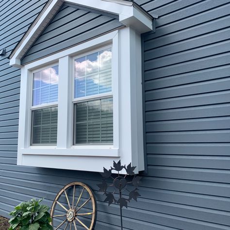 The photo is a close up on a home showing it's greyish-blue vinyl siding with large center window that has white trim. There's also a wagon wheel as decor against the home, and a large green plant, along with a windmill. In the reflection of the window, you can see the clear sky and fluffy clouds. Can You Paint Vinyl Siding On A House, How To Paint Vinyl Siding On A House, Outside Vinyl Siding Colors, Painted Vinyl Siding House Exteriors, Can You Paint Siding On A House, Updating Vinyl Siding Exterior, Can Vinyl Siding Be Painted, Painting Vinyl Siding House Exteriors, Painting Exterior Vinyl Siding