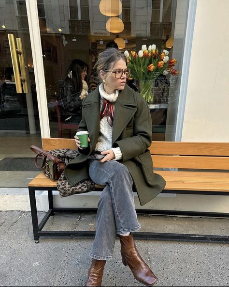 Parisian blond girl sitting on a bench with her coffee in hand. She is very in trend dressed. Her hair is up and is wearing brown squared glasses. She is wearing a thick white knitted turtleneck which she tied a thin elegant red satin  scarf around her neck. She associated this with grey jeans, brown leather block heels boots and a green jacket. Europe Outfits Cold, Casual Europe Outfits, Parisian Chic Style Fall, Khaki Jacket Outfit, Aesthetic Outfits Jeans, Winter Europe Travel, Winter Jeans Outfit, Green Coat Outfit, Outfits Black Jeans