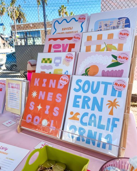 Colorful art prints on tiered display stand at Artist and Fleas fair in Venice, California. Shop these prints and many other vibrant pieces at nixieincolor.com. Display Art At Craft Fair, How To Display Art At A Craft Fair, Art Pop Up Display, Artist Market Display, Greeting Card Display Ideas, Card Display Diy, Pop Up Market Display Ideas, Art Market Display, Market Displays Booth Ideas