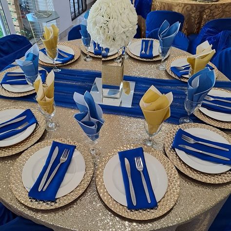 Take your table setup to the next level by using a gold sequin tablecloth paired with a royal blue runner that effortlessly exudes charm. Craft a captivating centerpiece by arranging flowers in an elegant vase and use a square mirror as your centerpiece base for a stunning appeal. Introduce gold chargers and white plates with royal blue napkins and silver cutlery for added allure. Also, add royal blue covers to your chairs to complete this setup. Royal Blue And Gold Table Decorations, Royal Blue Napkins, Mirror Chair, Gold Table Decorations, Royal Blue Wedding Theme, Gold Sequin Tablecloth, Event Decor Ideas, Party Decorations Table, Arranging Flowers