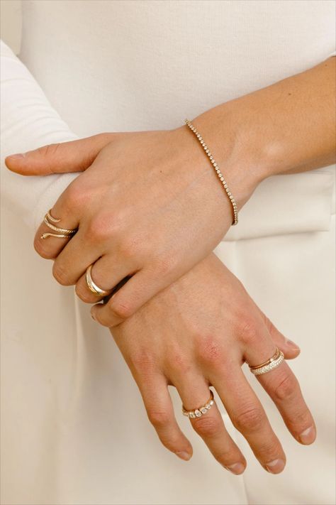 Two hands clasped together, the left wrist adorned with a delicate diamond bezel tennis bracelet, complemented by various gold rings on the fingers, including a snake ring, dome rings, and a diamond dome ring, set against a soft white fabric background. Modern Wardrobe, Bezel Diamond, Tennis Bracelet, Bezel Setting, Handmade Bracelets, Bend, Natural Diamonds, Gold Filled, Oregon