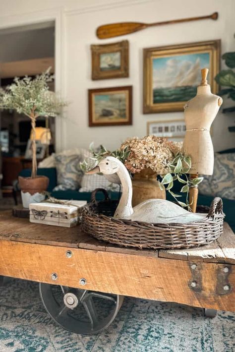 Embrace the rustic charm of winter with this enchanting coffee table centerpiece. A vintage wooden cart overflows with a cozy basket housing a faux goose nestled amidst a wintry floral arrangement of dried hydrangeas and flowing greenery. Add a faux topiary and a bundle of books for a touch of magic that spells pure winter bliss. French Country Coffee Table Decor, French Country Coffee Table, Faux Topiary, Christmas Coffee Table, Coffee Table Centerpiece, Basket Arrangement, Christmas Coffee Table Decor, Winter Floral Arrangements, Wooden Cart