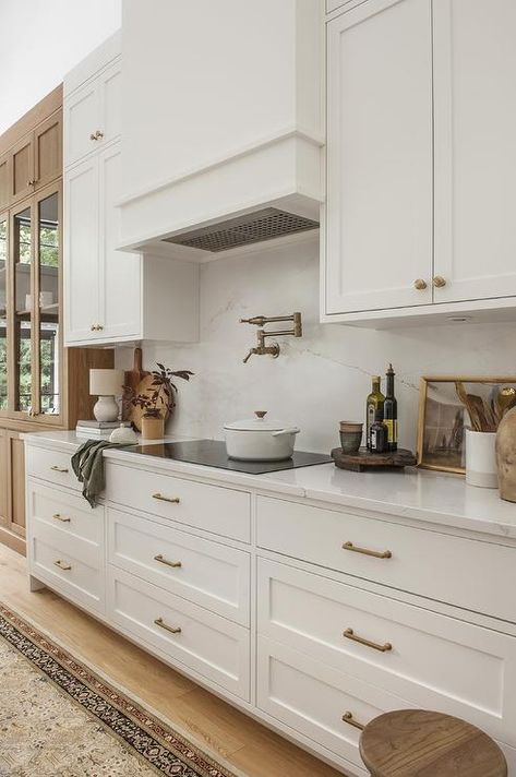 White and gold kitchen boasts a white range hood mounted against a marble-look slab backsplash between white inset cabinets with brass knobs. White And Gold Kitchen, Slab Backsplash, Kitchen With Marble, Classical Kitchen, Kitchen Hood Design, White Shaker Kitchen Cabinets, Off White Kitchens, Elegant Kitchen Design, White Shaker Kitchen