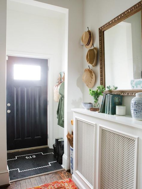 A vintage gold mirror hangs above an entry table created from the custom radiator cover in this eclectic home’s entryway. Pops of color are added with the vibrant antique rug and accessories while the black door and custom black and white tile work add sophistication to the space. Wall Heater Cover, Eclectic Entryway, Custom Radiator Covers, Contemporary Radiators, Hallway Ideas Diy, Vintage Gold Mirror, Heater Cover, Entryway Mirror, Hallway Designs