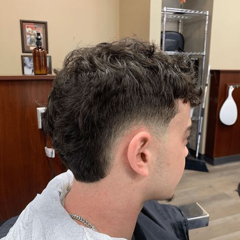 Side profile of a man with a Low Burst Fade haircut, showcasing a subtle and sophisticated fade that starts just above the ears. The hairstyle features thick, wavy hair on top, gradually tapering down to shorter lengths at the sides and back. This low burst fade provides a clean and polished look, suitable for both professional and casual settings. The background includes a barber shop setting with wooden panels and grooming tools, emphasizing the fresh, well-groomed style. Low Burst Fade, Burst Fade Haircut, Burst Fade, Man Beard, Thick Wavy Hair, Fade Haircuts, Short Haircut, Side Profile, Fade Haircut