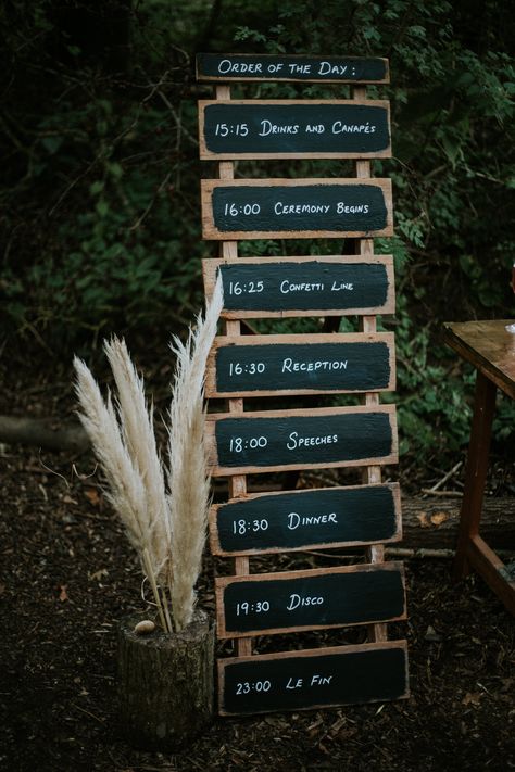 This shows an outdoor wedding. The focus is on a 'order of the day' sign in a rustic wooden black board sign. Next to it are some dried pampas leaves displayed on a trunk of wood. Order Of Service Wedding Sign, Blackboard Sign, Order Of The Day Sign, Dyi Wedding, Wedding Guest Signing, Lloyd Jones, Wood Photography, Plan A Wedding, Spa Ideas