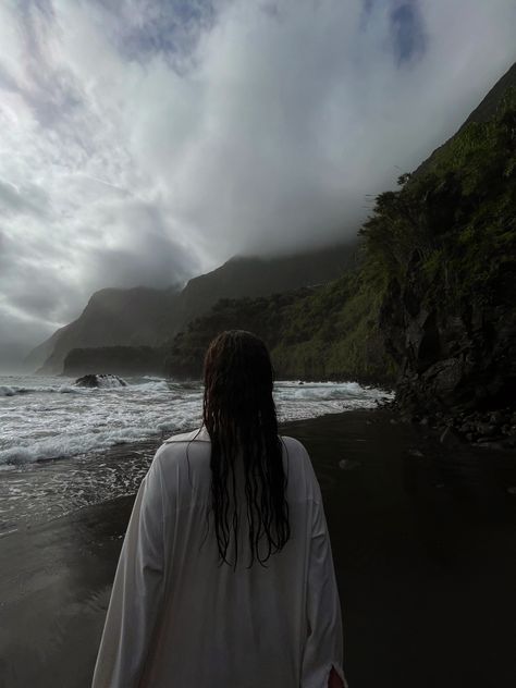 A picture of a girl on the Seixal beach on Madeira. Her wet hair from the sea is falling down onto her white shirt. She is facing the sea, not the camera. The sky is cloudy without the sun brightening it up, so the colours seem dark. Black Sand Beach Photo Ideas, Black Sand Beach Aesthetic, Seixal Beach Madeira, Madeira Outfit Ideas, Cloudy Beach Photoshoot, Cloudy Beach Aesthetic, Madeira Portugal Aesthetic, Wet Hair Aesthetic, Cloudy Photoshoot