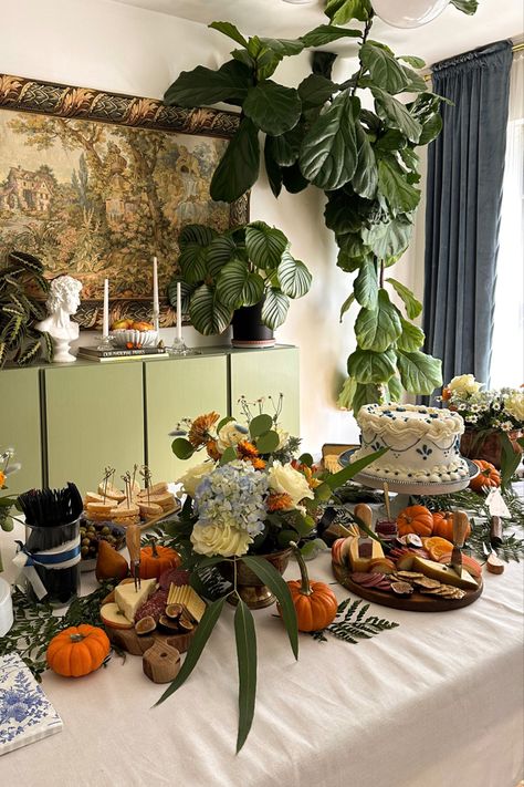 An elegant tablescape featuring a vintage-style blue and white Lambeth cake at its center, surrounded by blue and white platters. Mini pumpkins, rustic wooden charcuterie boards, and vibrant fall florals adorn the table. A tapestry hangs on the wall as a backdrop, and white taper candlesticks provide a warm, inviting glow. A harmonious blend of classic and autumnal elements Bib Decorating Station, Copper Containers, Lambeth Cake, Pimento Cheese Sandwiches, White Platter, Rustic Backdrop, White Tablecloth, Copper Vase, Tablescape Inspiration