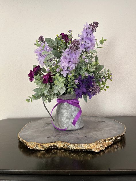 Transform your space into floral paradise with this exquisite roses and lilac flower arrangement in stunning shades of purple and lavender. This beautiful centerpiece features a blend of artificial miniature roses, lilacs, and eucalyptus for that touch of elegance. You will find your arrangement in a charming galvanized watering can or wash tub with wooden handles that adds that rustic country flair to any decor. Then it is topped with the finishing touch of a beautiful tied purple bow that will Simple Purple Table Decorations, Wisteria Flower Centerpieces, Dark Purple Floral Arrangements, Purple Floral Party Decor, Purple And Green Wedding Centerpieces, Purple Centerpiece Ideas, Lilac Centerpieces, Church Tables, Lavender Decorations