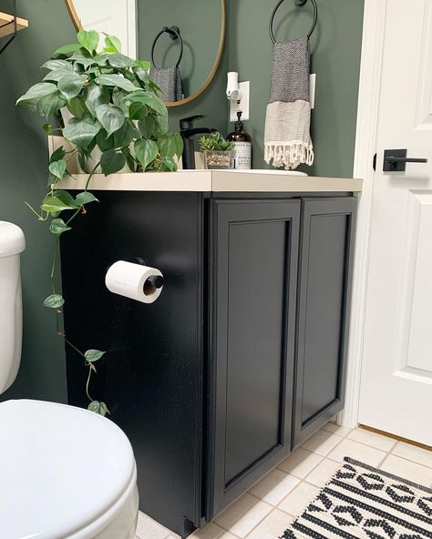 This bathroom is designed with a black-painted vanity and a light beige countertop. Sage green walls give the room a modern look, along with a round, gold mirror hung above the sink. A striped gray hand towel and potted plant accessorize the space. Black Vanity Green Walls, Black Vanity Gray Walls, Dark Green Bathroom Black Vanity, Bathroom Paint With Dark Cabinets, Black Cabinet Green Wall Bathroom, Black Vanity Green Walls Bathroom, Dark Vanity Small Bathroom, Green In Bathroom Ideas, Green Bathroom With Black Vanity