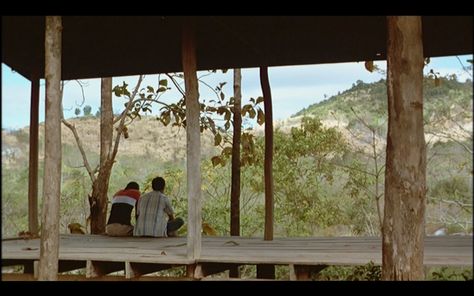 Tropical Malady Weerasethakul places the characters in the middle ground, facing away from us. Frames within frames and the guiding lines of the wooden structure help guide the eye to the two lovers. The idyllic Thai countryside in the extreme background completes the image. Frames Within Frames, Film Stills Nature, Movie Landscapes Scene, Thai Countryside, Cinematic Field Photography, Farm Cinematography, Tropical Malady, Movie Composition Cinematography, Herd Of Elephants