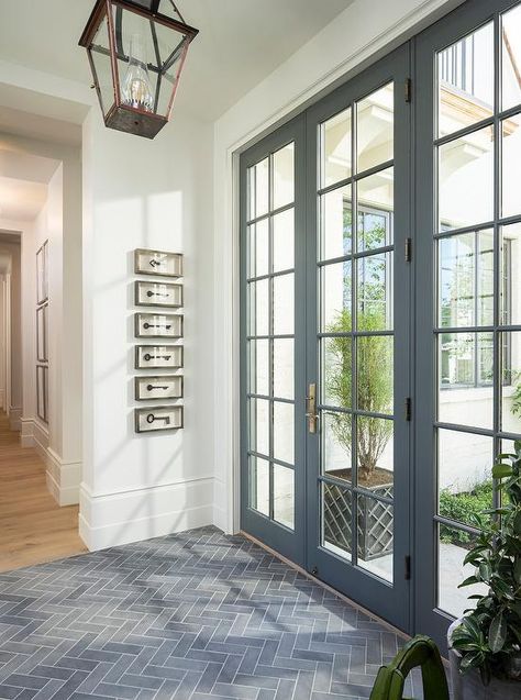 Gray slate herringbone tiles lead to a a gray glass paned front door illuminated by a bronze carriage lantern while framed vintage keys in shadow boxes are mounted the white wall of this stunning gray and white foyer. White Foyer, Entryway Tile, Foyer Flooring, Entry Tile, Entryway Flooring, Entryway Inspiration, Entryway Lighting, Herringbone Floor, Foyer Decorating