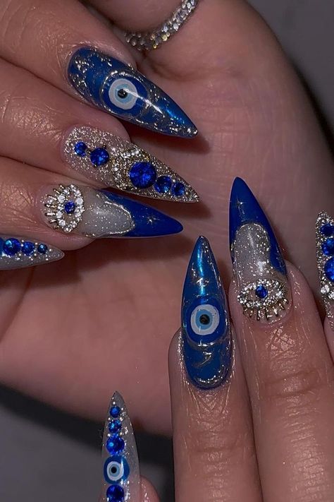 These stiletto nails are a bold statement of glamour and mystique. The rich blue base is graced with a protective Evil Eye design, bringing a touch of tradition to a modern look. Alternating nails are encrusted with a mix of glittering silver and sapphire-hued rhinestones, creating a luxurious sparkle that's sure to capture the eye. The eye-catching combination of intricate details and dazzling embellishments makes these nails an enchanting choice for anyone looking to add a dramatic flair to their style. ✨  // Photo Credit: Instagram @socialclimbernail Evil Eye Inspired Nails, Evil Eye Nail Design, Dark Blue Evil Eye Nails, Nails 2023 Trends Evil Eye, Turkish Eye Acrylic Nails, Evil Eye Nail, Blue Evil Eye Nails, Press On Nails Evil Eye, Evil Eye Manicure Nail Designs