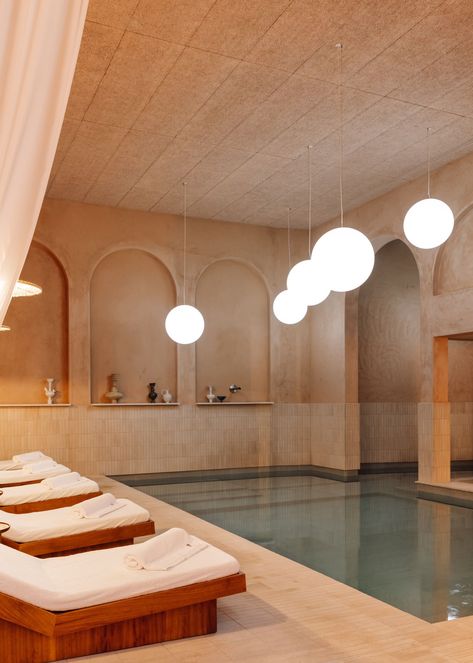 Inspired by ancient Roman thermal baths, the Spa at The Rebello is centered around serenity, calmness, and luxury, with a strong focus on wellness and relaxation. The centerpiece of the spa is an indoor pool lined with studio-designed iroko daybeds and billowing drapery. The pool is enhanced by programmed lamps that mimic the natural phases of the sun and moon, including dawn, sunset, and moonlight. Indoor Pool And Sauna, Indoor Pool Lighting, Pool House Lighting, Massage Room Lighting, Cold Plunge Spa, Interior Pool Design, Magnesium Pool, Wellness Spa Design, Hotel Pool Design