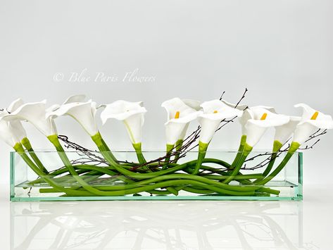 "Modern Long White REAL TOUCH Calla Lily Arrangement Artificial Faux Centerpiece Floral Flower Table Decor Silk Flowers Glass Vase French  * Arrangement comes in a 12\" or 24\" clear vase. (24\" arrangement shown on images)  * Feel free to contact us if you need more information about the arrangement. * At first look, people might not believe this is a permanent botanical design! * The arrangement contains the finest faux white calla lilies in a long rectangular glass vase. * The flowers have larger heads and they feel soft and lovely, real, and lively.  * All are arranged in a very modern and elegant long rectangular vase. * Perfect centerpiece on a long or rectangular dining table.  At Blue Paris Flowers we take pride in producing unique and luxurious floral designs. We handcraft each it Calla Lily Arrangement, Flowers Glass Vase, Flower Table Decor, Lily Arrangement, Lily Centerpieces, Paris Flowers, Rectangular Vase, Corporate Flowers, Flower Table