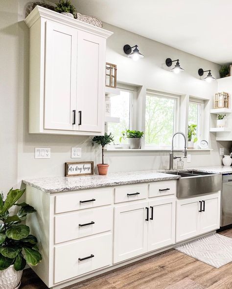 These white cabinets with black hardware are topped by a gray and black countertop in this farmhouse kitchen. A stainless apron sink is fitted into the counter under white windows. There is no backsplash behind the sink. Kitchen Ideas Marble Countertops, White Kitchen Countertop Ideas, White Dream Kitchen, Over Kitchen Sink Lighting, White Tile Kitchen Backsplash, Kitchen Sink Lighting, White Tile Backsplash, Organizing Hacks, Farmhouse Kitchen Design