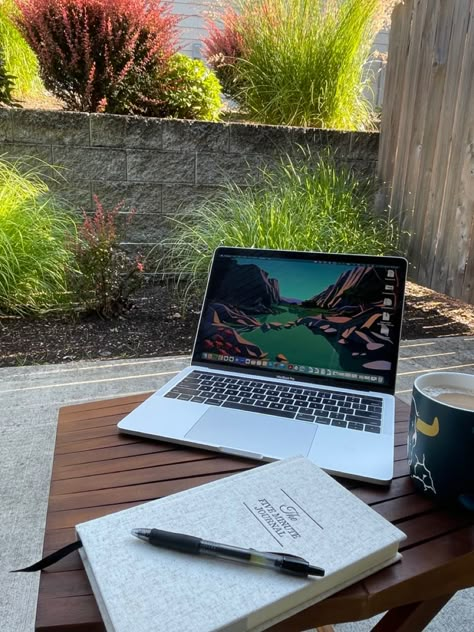 A laptop, mug of coffee, and a journal are sitting on an outdoor table on a patio. The patio is surrounded by bushes and plants. Outdoor Office Space Patio, Work At Home Aesthetic, Wfh Inspiration, Work Astethic, Working From Home Aesthetic, Outdoor Office Space, Remote Work Aesthetic, Wfh Aesthetic, Work From Home Desk Setup