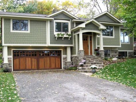 This lovely split-level home blends in beautifully with the trees and lawn around it, thanks to its soft gray-green siding. The stone facade and steps provide a touch of nature while the carriage-style garage doors and elegant wood front door add a bit of Craftsman charm. Split Level House Exterior, Split Level Exterior, Green Siding, Split Level Remodel, Split Foyer, Exterior House Remodel, Ranch Remodel, Stone Steps, Split Level House