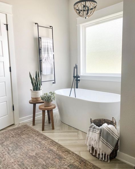 A wood-beaded chandelier hangs above a white freestanding bath with a black detachable shower head. Two wood stools with planters sit nearby along a herringbone stone tile floor and french country bath rug. A woven basket and steel wall-mounted towel rack hold farmhouse tassel towels. Stand Alone Bath Tub, Bathroom Towel Rack Ideas, Standing Bathtub, Standing Tub, Stand Alone Tub, Bathtub Decor, Bathtub Walls, Creative Bathroom, Modern Farmhouse Bathroom