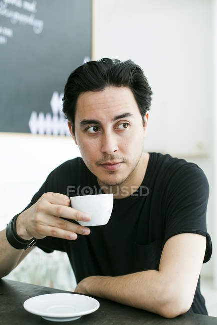 Man holding coffee cup while sitting at table — indoors, 30 34 years - Stock Photo | #151412326 Someone Holding A Mug Reference, Someone Sitting At A Table Reference, Sitting At The Table Pose, Person Holding Coffee Reference, Arm On Table Reference, Person Holding Mug Reference, Leaning On Table Reference, Person Holding Cup Reference, Holding Coffee Pose Reference