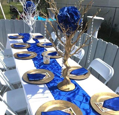 Compliment the liveliness of an outdoor setting with a bright and baroque table décor. Dress up your table with a white tablecloth and line with a ravishing royal blue taffeta runner. Treat your guests with a delicate plate setting using gold chargers and royal blue napkins. Create an alluring focal point using gold and white Manzanita trees centerpieces embellished with royal blue flower balls and elegant beaded chains. Complete the look by placing your luminaires in crystal candle holders. Royal Blue Centerpieces, Royal Blue Wedding Decorations, Quince Centerpieces, Manzanita Centerpiece, Royal Blue Wedding Theme, Royal Blue Quince, Blue Table Settings, White And Gold Decor, Blue Wedding Decorations