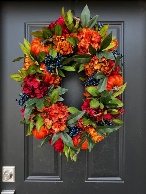 This Oval Bountiful Harvest Wreath brings the warmth and richness of the season straight to your door. Lush, vibrant hues of burnt orange, deep reds, and golden yellows are interwoven with lifelike pumpkins, bold peonies, and clusters of deep blue berries. The oval shape adds an elegant, unexpected twist to the traditional round wreath, making it a striking statement piece. Whether hung on your covered front door or displayed indoors, this handcrafted wreath is perfect for welcoming fall and cel Wreath For Office Door, Tombstone Flowers, Centerpiece Craft, Floral Door Wreaths, Blue Berries, Door Wreaths Diy, Harvest Wreath, Wreath Hanger, Bountiful Harvest