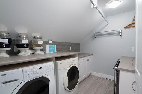 An attic laundry room features white cabinetry paired with gray countertops and a gray glass tiled backsplash placed under a sloped ceiling. Gray Countertops, Farmhouse Laundry Room Ideas, Glass Closet Doors, Attic Renovation Ideas, Glass Closet, Attic Closet, Attic Playroom, Attic House, Small Attic