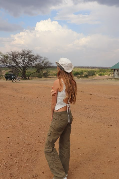 A blond girl looks at the Savana. She's dressed in a white laced tank top and green cargo pants, her cowboy hat puts the outfit together. African Trip Outfits, Safari Aesthetic Outfit, Safari Outfit Women Africa, Tanzania Outfit, Safari Chic Outfits, Kenya Outfits, Safari Fits, South Africa Outfits, Safari Outfit Women