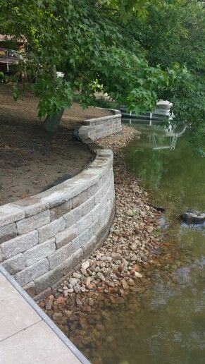 We tore out an old wooden reatining wall and replaced with a tumbled retaining wall block. We placed creek rock on the front side of the wall for a visual apperance and to also hide the concrete footing. We also extended the paver walkway, attached the dock, and added topsoil to freshen everything up. Located in central MO.  ** www.greenacresmidmo.com *** Creek Retaining Wall, Pond Retaining Wall Ideas, Lake Retaining Wall Ideas, Seawall Ideas, Lake Landscaping, Garden Decoration Ideas, Lake Dock, Paver Walkway, Landscaping Retaining Walls