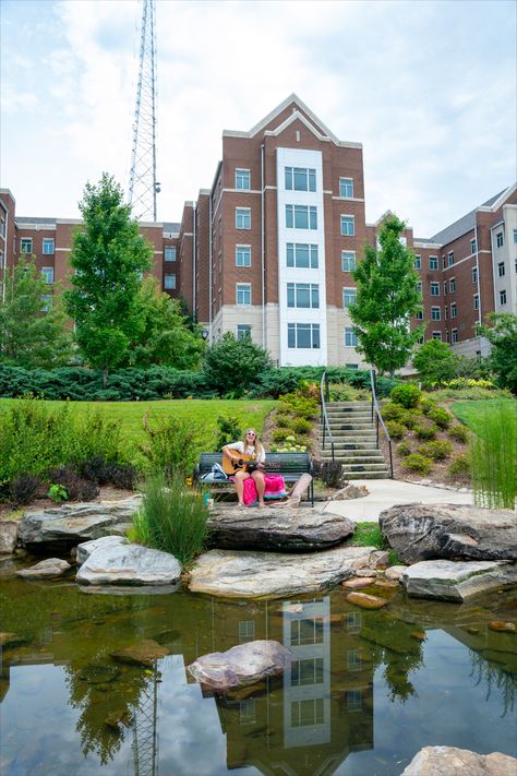 Belmont University Bear creek; pond with stones and muddy water. Female student sitting on a bench playing the acoustic guitar. Large dorm building in the background with blue sky and telephone pole. Belmont University Nashville, Nashville Aesthetic, Belmont University, College Tour, Visit Nashville, Spring Semester, Nashville Trip, Baylor University, Spring Photos