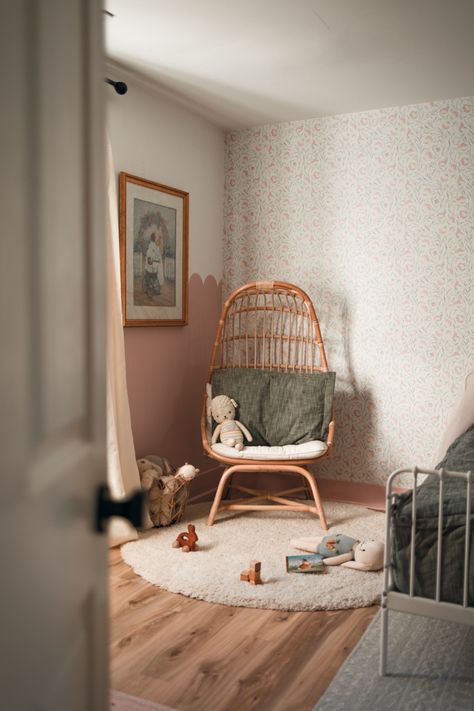 Photo features a open, the door peeking into a children’s bedroom with a rattan chair in a corner with toys on the floor. The corner of a bed is shown floral wallpaper on one wall and dusty rose pink scallop wall on the other wall with a vintage painting of two sisters under a floral arch on the scalloped wall. White rug on hardwood floor. Dusty Rose Toddler Room, Beige Nursery Girl, Toddler Bedroom Green, Bedroom Dusty Rose, Shared Toddler Room, Green Girls Room, Dusty Rose Nursery, Girls Bedroom Green, Floral Wallpaper Vintage