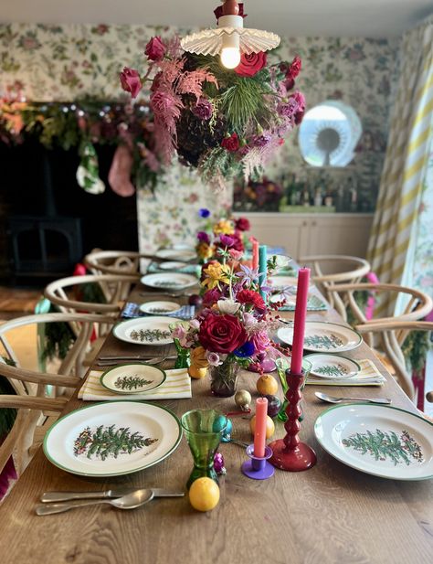 Colourful Christmas tablescape on large wooden table with Spode dinnerware which is white with a green rim and Christmas tree in the centre. Above the table hangs an extravagant foliage cloud filled with greenery, red roses and pink flowers. The centre of the table is fulled with mini floral displays. bright and colourful candlesticks with fruit and baubles scattered throughout. Eclectic Christmas Table Setting, Red Roses Christmas Table Decor, Pink Red Christmas Table, Colourful Candlesticks, Colourful Christmas Table, Spode Dinnerware, Modern Christmas Tablescape, Modern Christmas Table, Large Wooden Table