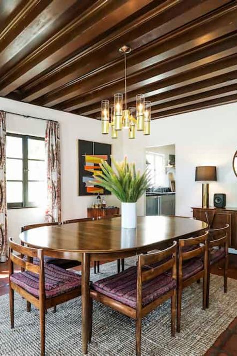 The dining room of his Spanish Colonial Hollywoodland home's focus is the oval-shaped wooden dining table set lighted by a gorgeous pendant light that hangs above exposed beams. The green indoor plants and colorful decor pop from the bright white wall background. Get more ideas on how to make a house to look like a celebrity home and see more of the interior and with this house tour. #diningroom #celebrityhomes #housetour   Courtesy of toptenrealestatedeals Dining Table Spanish, Dining Room Decor Oval Table, Oval Wooden Table, Spanish Dining Room Hacienda Style, Modern Mexican Dining Room, Spanish Revival Dining Room, Colonial Revival Dining Room, Dining Table Against Wall, Spanish Colonial Dining Room