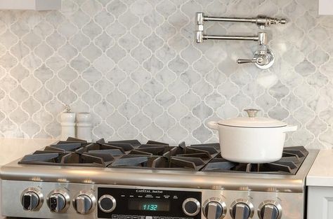 A stainless steel stove is mounted against gray marble arabesque cooktop tiles fitted with a polished nickel swing arm pot filler. Marble Arabesque Backsplash, Arabesque Tile Kitchen, Arabesque Tile Backsplash Kitchen, Cooktop Backsplash, Arabesque Tile Backsplash, Arabesque Backsplash, Backsplash Arabesque, Kitchen Backsplash Tile Designs, Marble Backsplash Kitchen