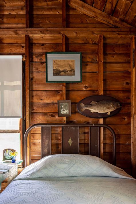 This cottage bedroom from the pages of The Maine House II book epitomizes the phrase 'quintessentially Maine'. Rough wood walls act as a backdrop to an eclectic collection of artwork—including a taxidermy fish, a yellowed landscape sketch, and a family photo— which are aptly mounted to exposed studs. A metal bed frame sets off a crisp white quilt and matching roman shade. Bedside accessories include a freestanding alarm clock and wildflowers. Rough Wood Walls, Maine Aesthetic Interior, Bedside Accessories, Large Wall Art Cabin Maine, Beachy Bedding Maine Cottage, Old Maine House, Maine Map Art, Bed Frame Sets, Blue Coffee Tables