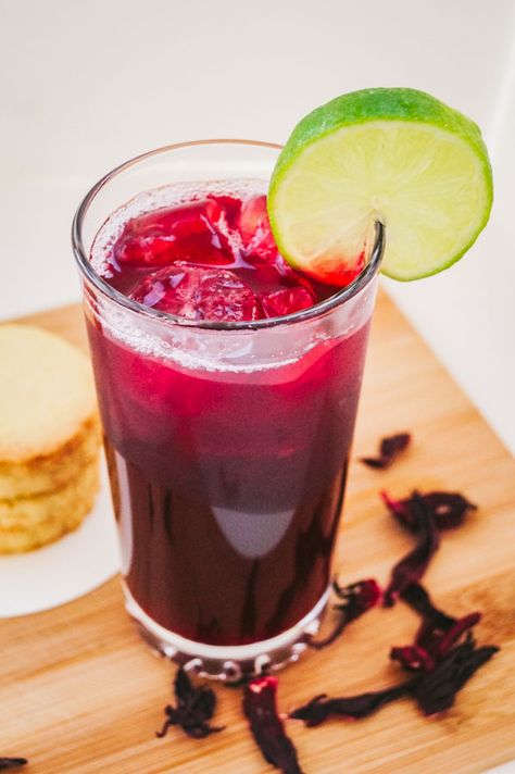 an angled overhead shot of zobo drink garnished with a slice of lime, beside it are stack of cookies Zobo Drink, Hibiscus Drink, Spiced Drinks, Nigerian Recipes, Orange Clove, Hibiscus Plant, Sparkling Drinks, Popular Drinks, Lime Soda