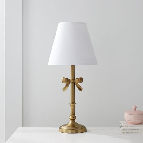 Tie their room together with this charming table lamp. It's expertly hand finished to emulate brushed gold and it comes topped with a white linen shade to cast soothing, filtered light around the room. DETAILS THAT MATTER Expertly crafted with aluminum and steel. Lamp base features a ribbon and is hand finished in a water-based Brushed Gold. On/off switch located on in-line; white cord. Shade is included and exterior is made from linen. Interior is lined with styrine. All our shades are designed Coquette Table Lamp, Gold Bow Lamp, White And Gold Table Lamp, Cute Gold Lamp, Silver And Gold Room Decor, Vintage Modern Room Decor, Gold And White Lamps, Pink Lamp Shades, White And Gold Lamps