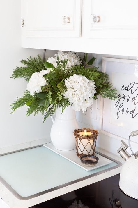 Winter white large head hydrangeas and greenery mixed in a white pitcher becomes cheery winter decor. Christmas Decor With Hydrangeas, Hydrangea Christmas Centerpiece, January Flower Arrangements, January Flower, Holiday Floral Arrangements, Winter Floral Arrangements, White Floral Arrangements, Flowers And Greenery, White Pitcher